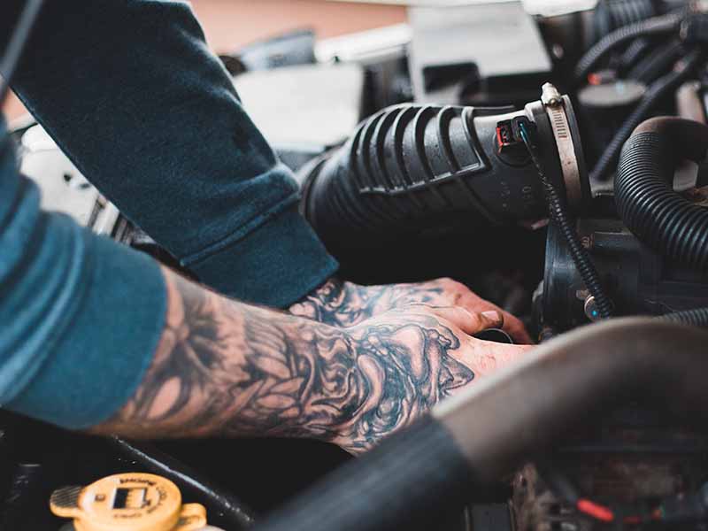 Man with tattooed hands and arms working on a car with the hood popped open.
