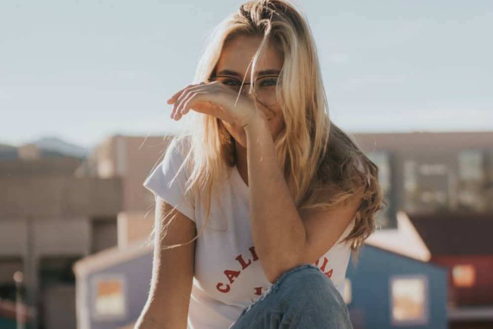 long-haired blonde woman with glasses in front of buildings